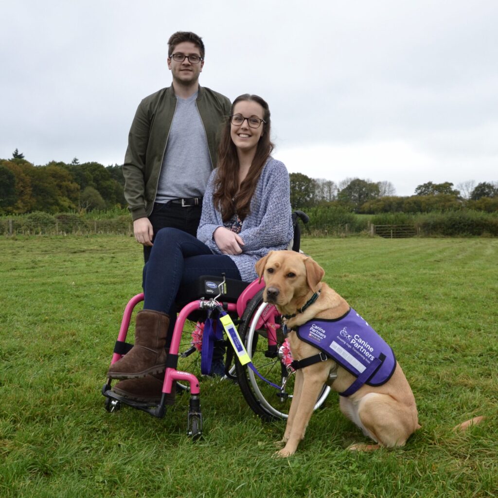 A white ambulant male and a white female using a pink wheelchair are outside with a red fox assistance dog.