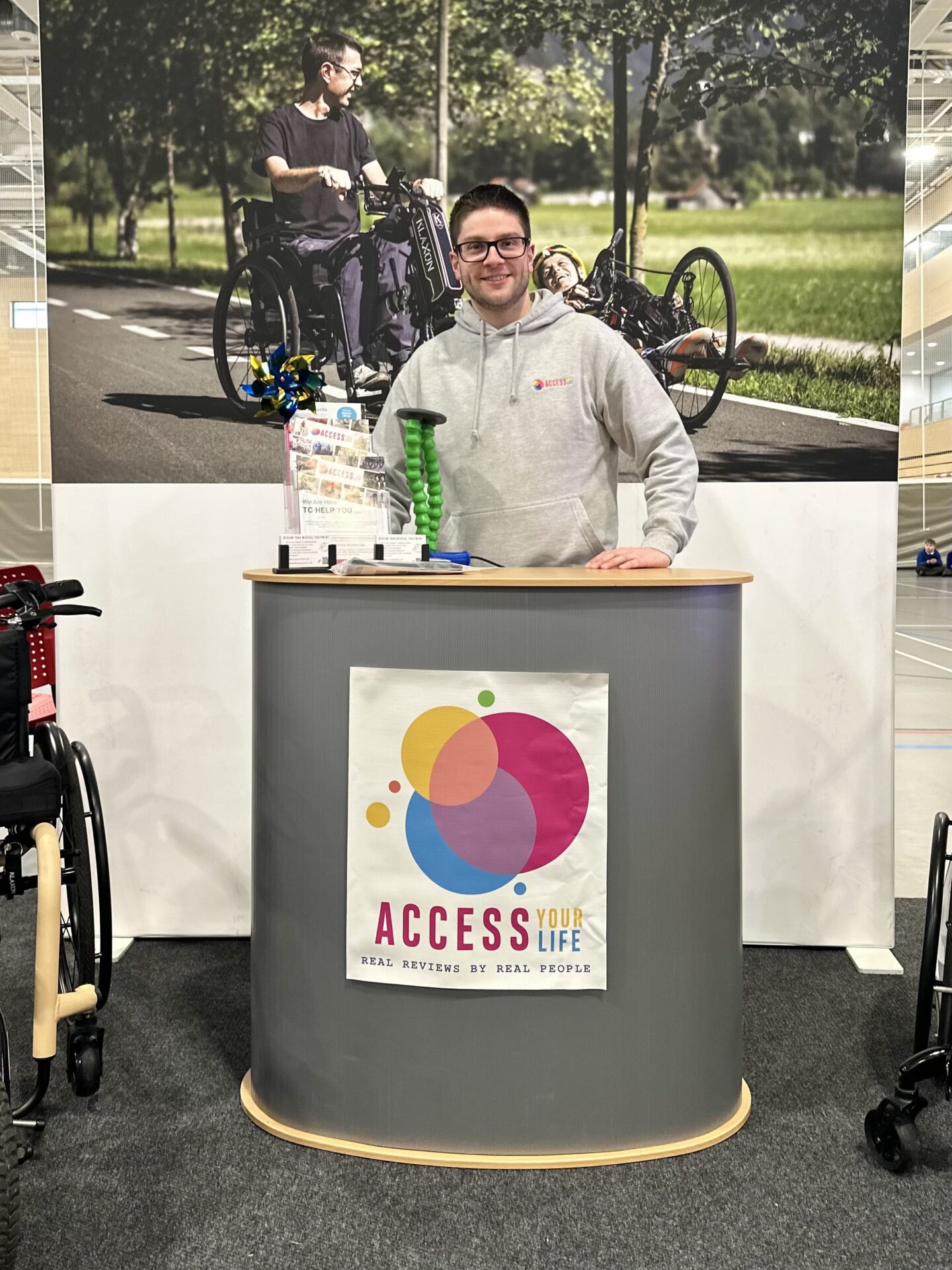 A white man with dark hair and glasses stood behind an exhibition stand with AYL's multicoloured logo on the front. A large photo of two individuals using mobility aids behind the the stand further underscores AYL's commitment to authentic disability advocacy.