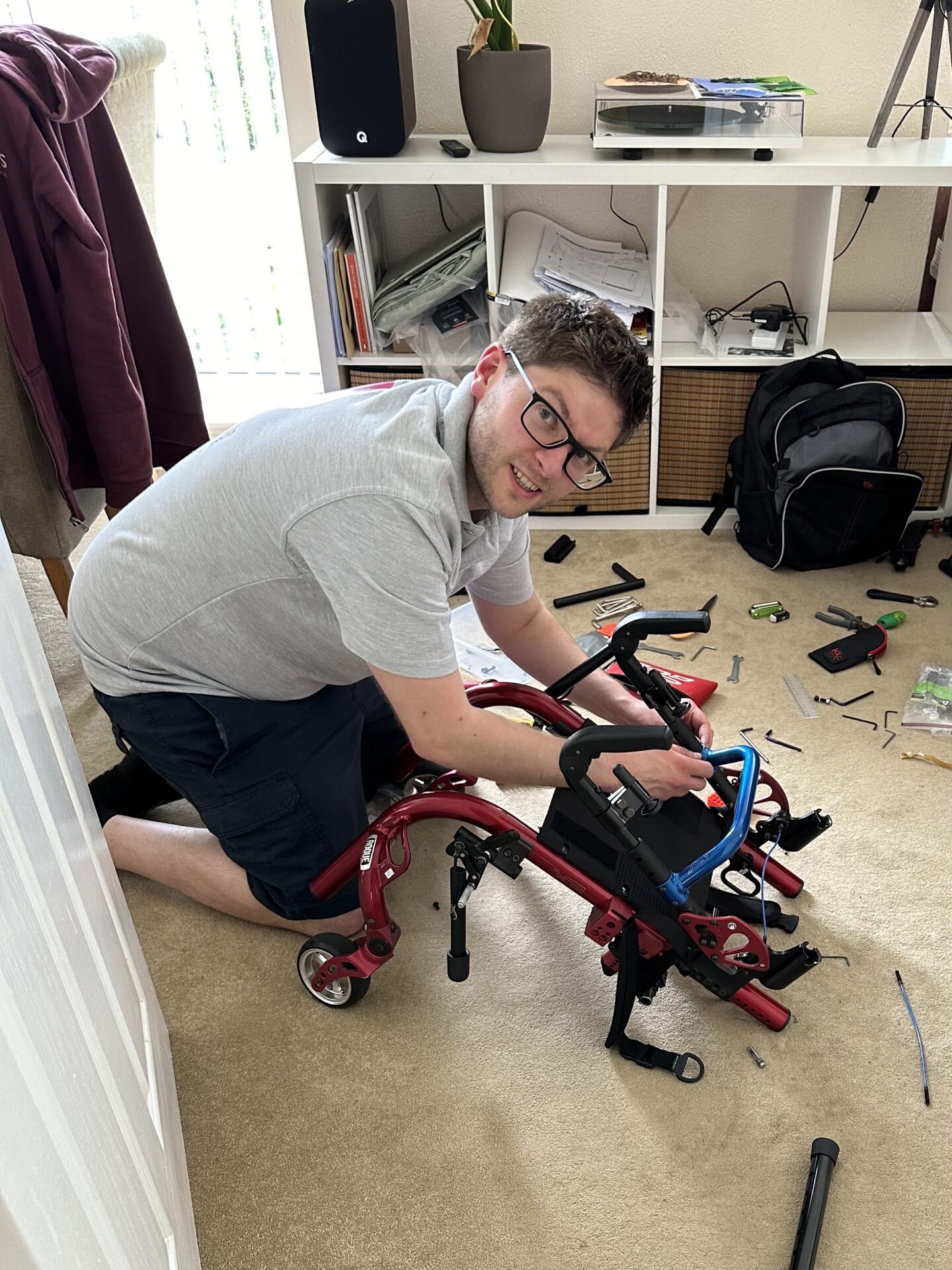 A white man with dark hair and glasses kneeling on the ground, fixing a red and blue paediatric manual wheelchair.