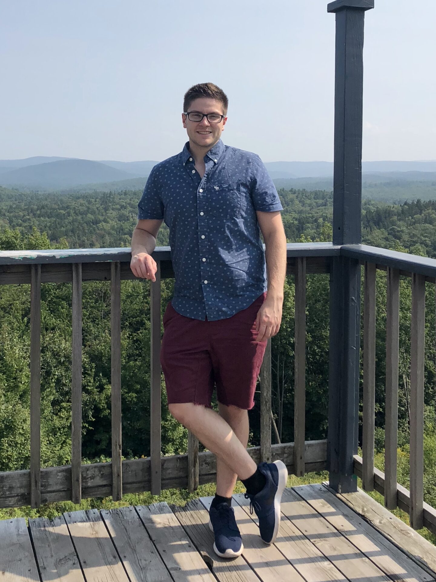 Photo of a man named Felix Ledger-Beadell, posing on a deck, overlooking a background shot of a forests stretching out into rolling hills that reach as far as the horizon.