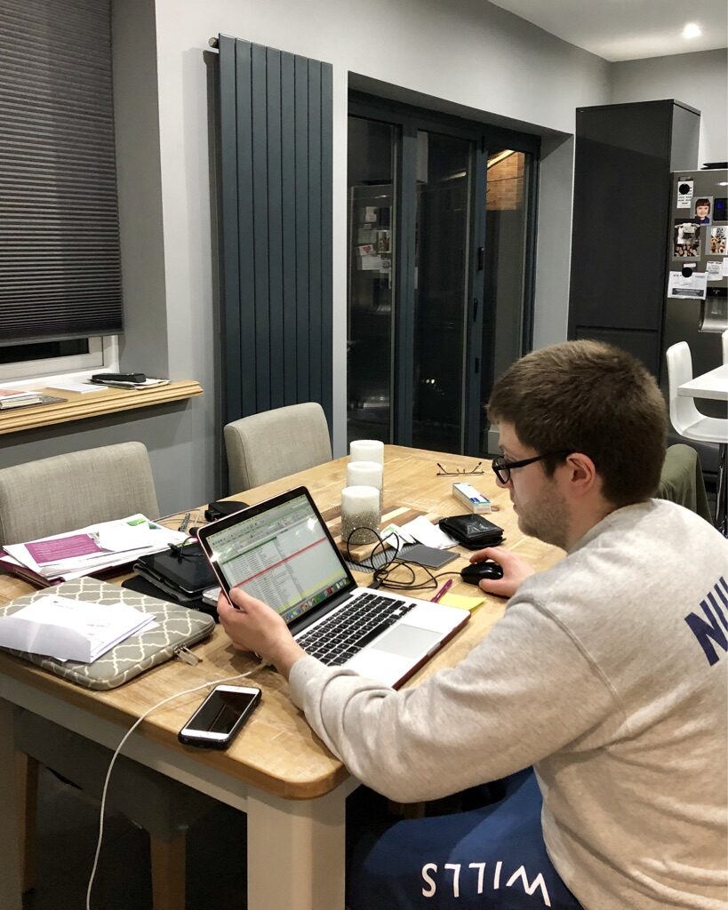 Photo of a bespectacled man with short brown hair sitting at a table, staring at a spreadsheet on a Laptop. A large variety of items are strewn across the table, suggesting the man has been working at it for some time.