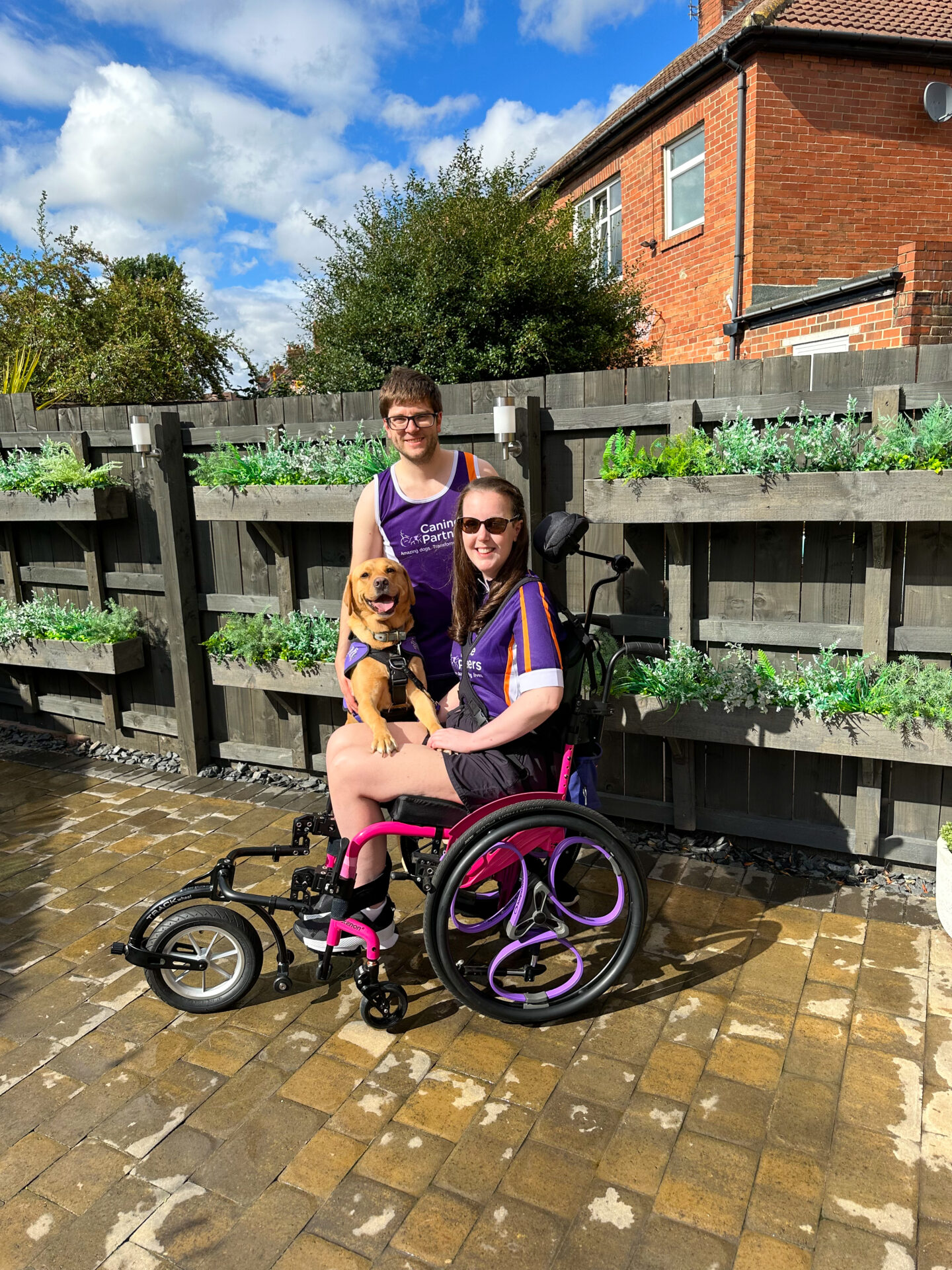 A white man and woman, both with dark hair and glasses, dressed in sports wear. The woman is using a pink manual wheelchair, with a 'Canine Partners' assistance dog (identifiable by its distinct purple coat) resting its front paws on the woman's lap.