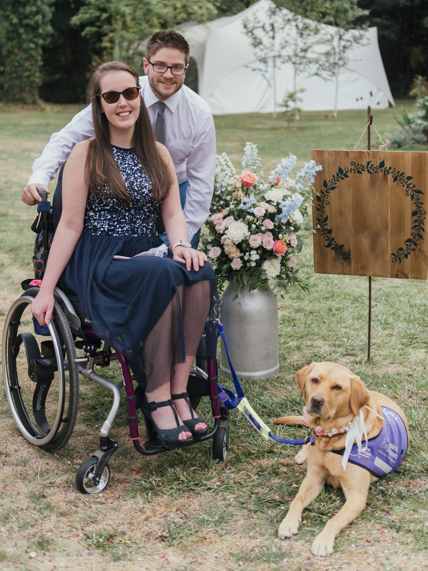 A white man and woman, both with dark hair and glasses, dressed in formal attire. The woman is using a purple manual wheelchair, with a 'Canine Partners' assistance dog (identifiable by its distinct purple coat) lying to the right of a large flower vase filled with many different coloured flowers, placed next to the couple.