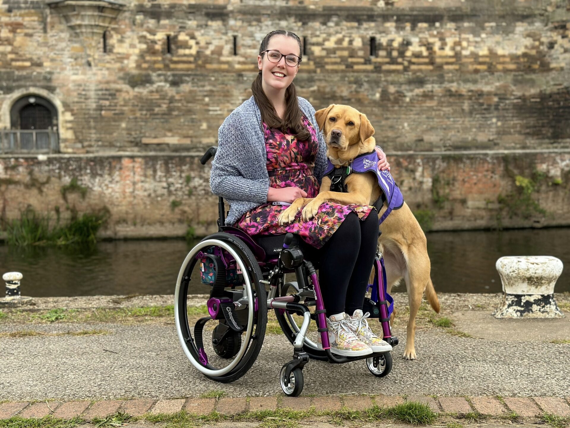 A white woman with brown hair and glasses, wearing a floral dress and sitting in a purple wheelchair, with a 'Canine Partners' assistance dog (identifiable by its distinct purple coat) resting its front paws on her lap.