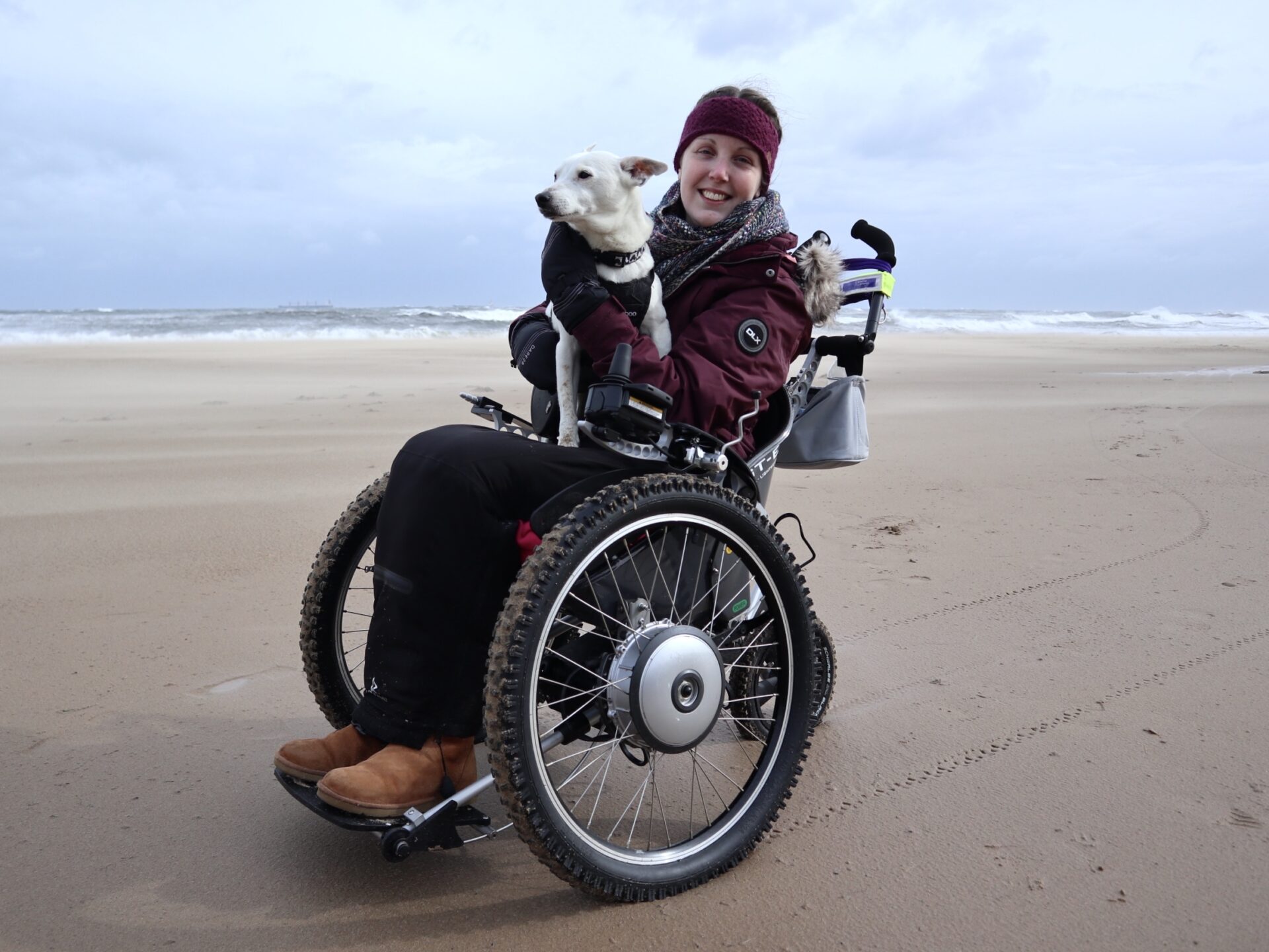A white female wearing winter clothing, sitting in a grey all-terrain powered wheelchair, with a white dog sitting on her lap. They're on the beach.