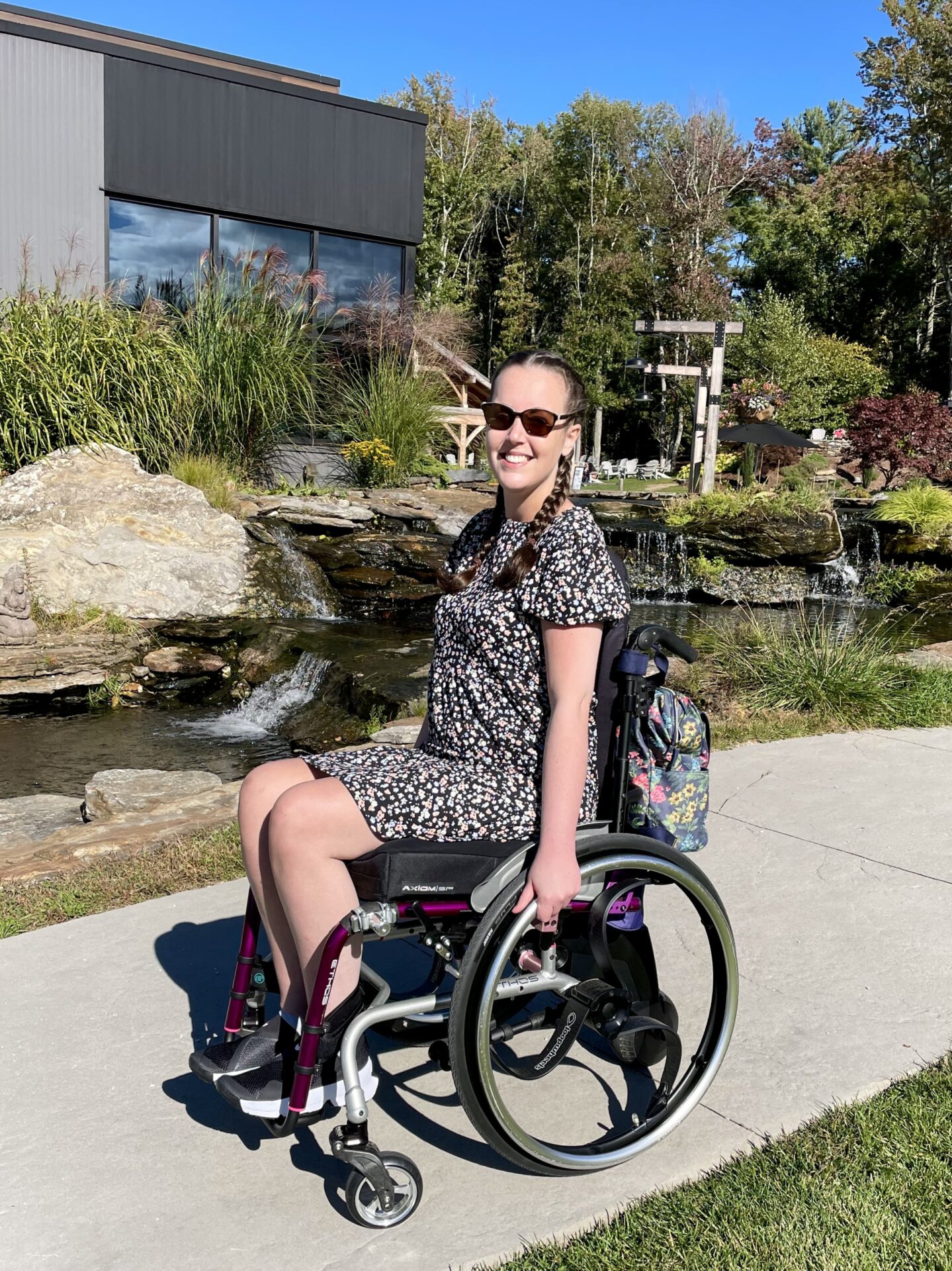 A white woman with brown hair and sunglasses, wearing a floral dress and sitting in a purple wheelchair, with a waterfall in the background.