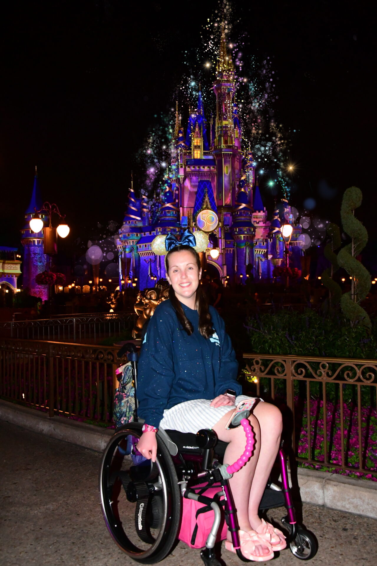 A white woman with brown hair and glasses, wearing a stripy dress and sitting in a purple wheelchair, with the Walt Disney World castle in the background.