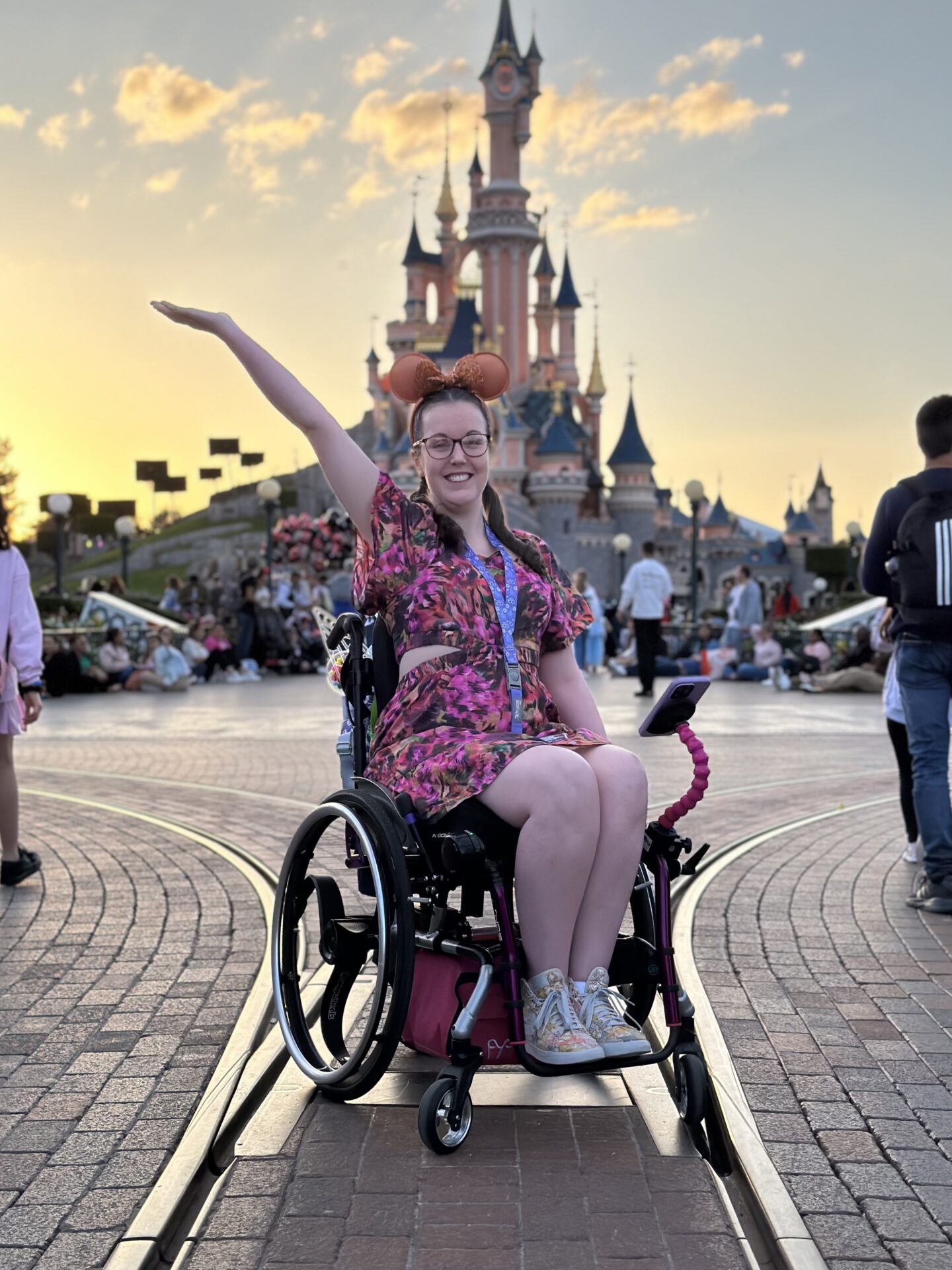 A white woman with brown hair and glasses, wearing a floral dress and sitting in a purple wheelchair, with the Disneyland Paris castle in the background.