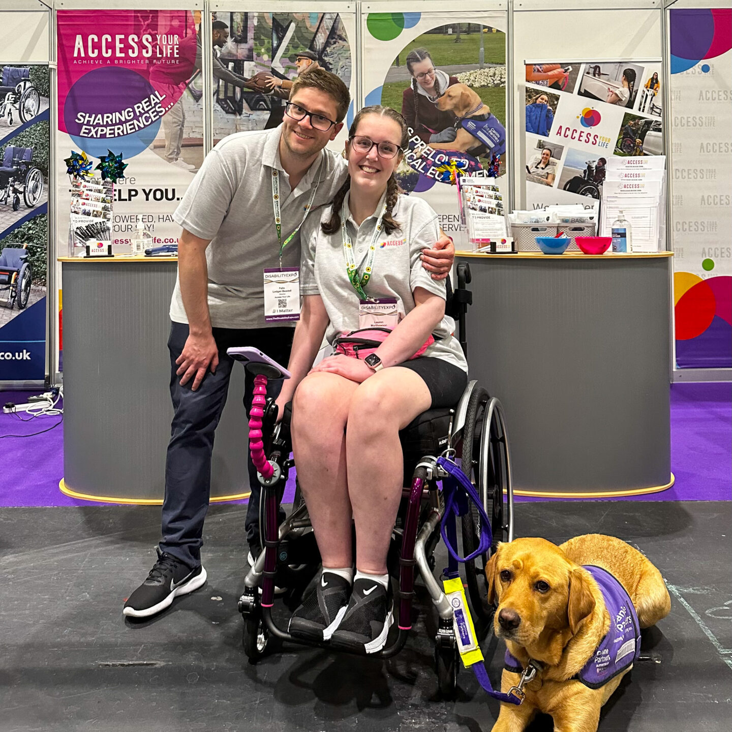 A white man and woman, both with dark hair and glasses, in front of a multicoloured exhibition stand. The woman is using a purple manual wheelchair, with a fox red assistance dog lying alongside.