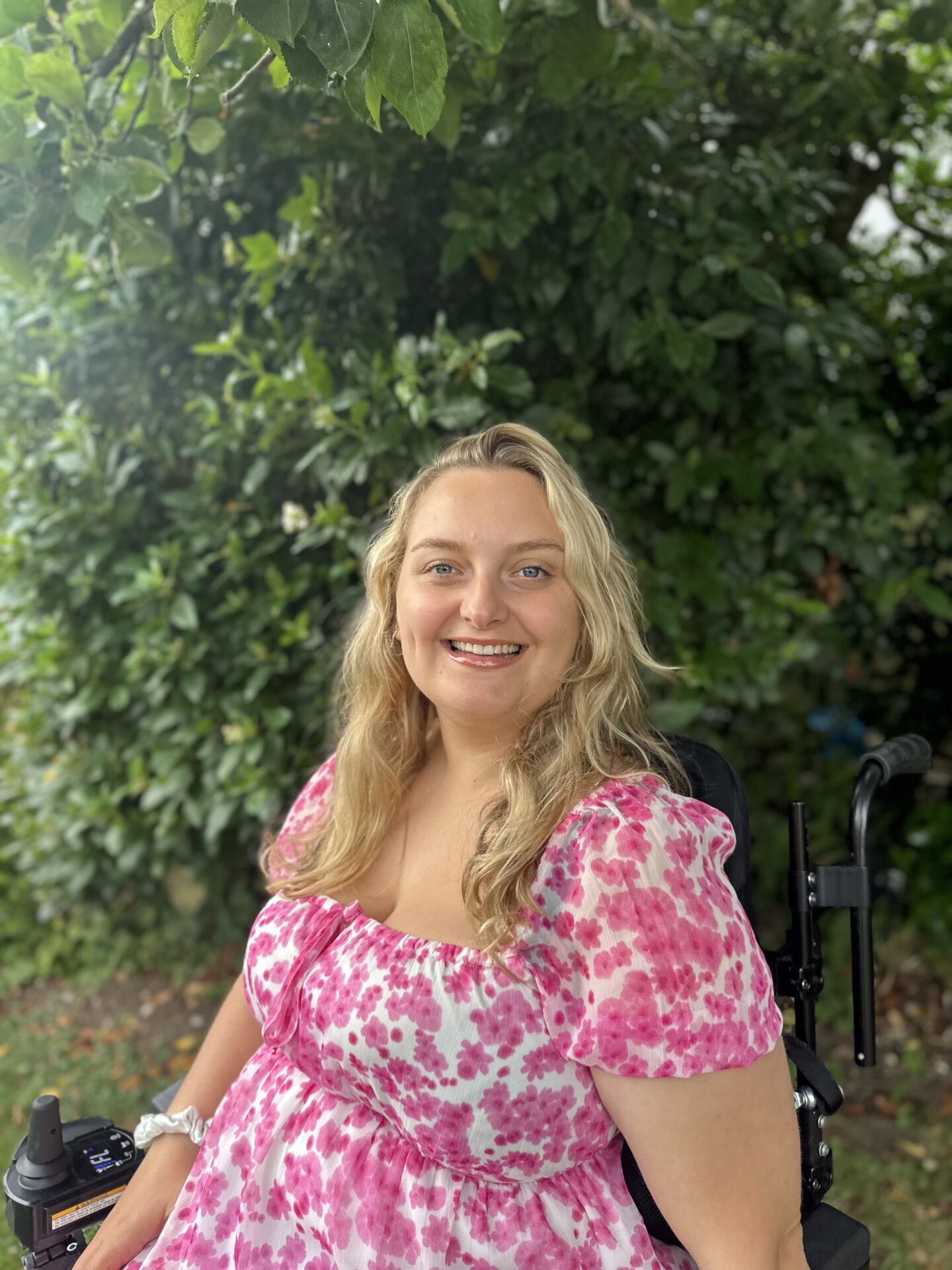 Profile photo of a blonde haired, smiling women wearing a pink dress, using an electric wheelchair. In the background, flowering tree branches can be seen.