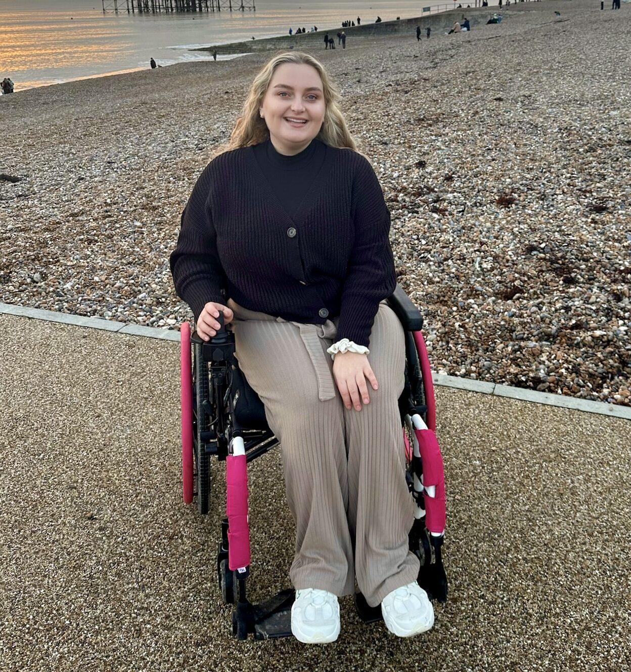 Profile photo of a woman using a self-propelling wheelchair with pink highlights on the push rims & castor legs, posing for a photo on a pebbly beach, with the water's edge just visible in the background.