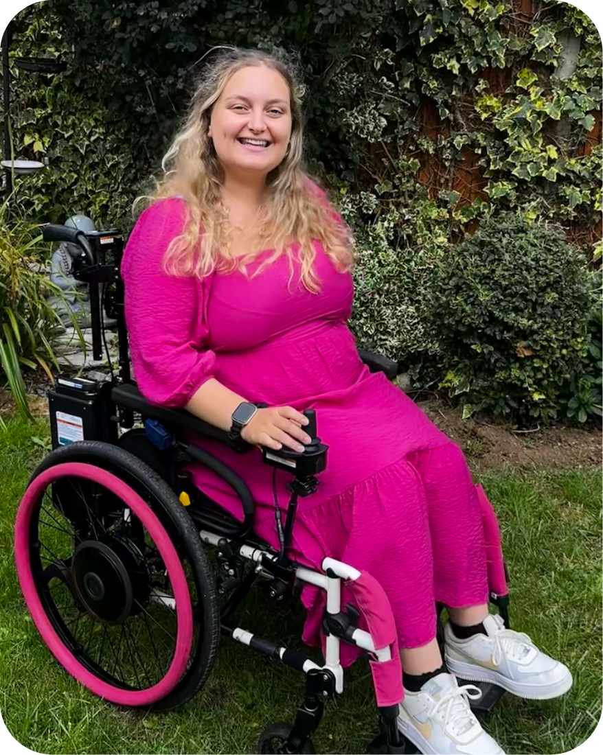 Photo of a smiling, blonde haired woman in an electric wheelchair. The woman is wearing a pink dress, pink sleeves, & matching pink colouring on her wheelchairs rims, with off colour white shoes. In the background, a garden & a statue of the Buddha can...