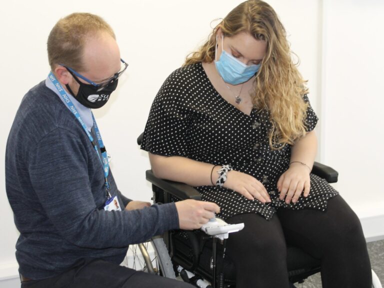 Picture of a blonde woman in an electric wheelchair, & a bespectacled man, with a receding blonde hairline, examining the joystick of the wheelchair. Both are wearing facemasks, & the man is wearing a NHS branded lanyard, from which we can infer he is some sort of medical professional.