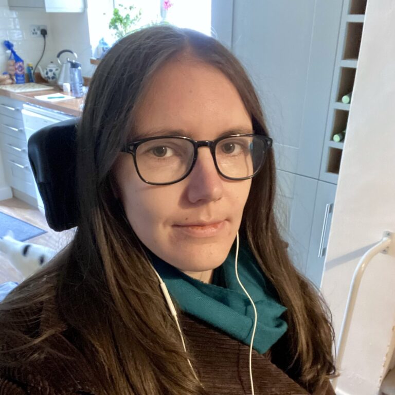 Selfie photo of a brunette women, wearing a blue scarf, glasses, & wired earbuds. From the headrest behind her we can infer that she is sitting down, & from the window sill and the work surfaces in the background we can infer this photo was taken in a kitchen.