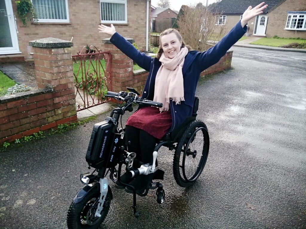 Photo of a brunette woman posing in a wheelchair with a 'Rehasense PAWS (Power Assisted Wheelchair System)' bike style front power add-on attached. The woman is wearing a pink scarf, blue coat, red skirt & black leggings, & is raising her arms out into the air, demonstrating the wheelchair + bike style add-on attached.