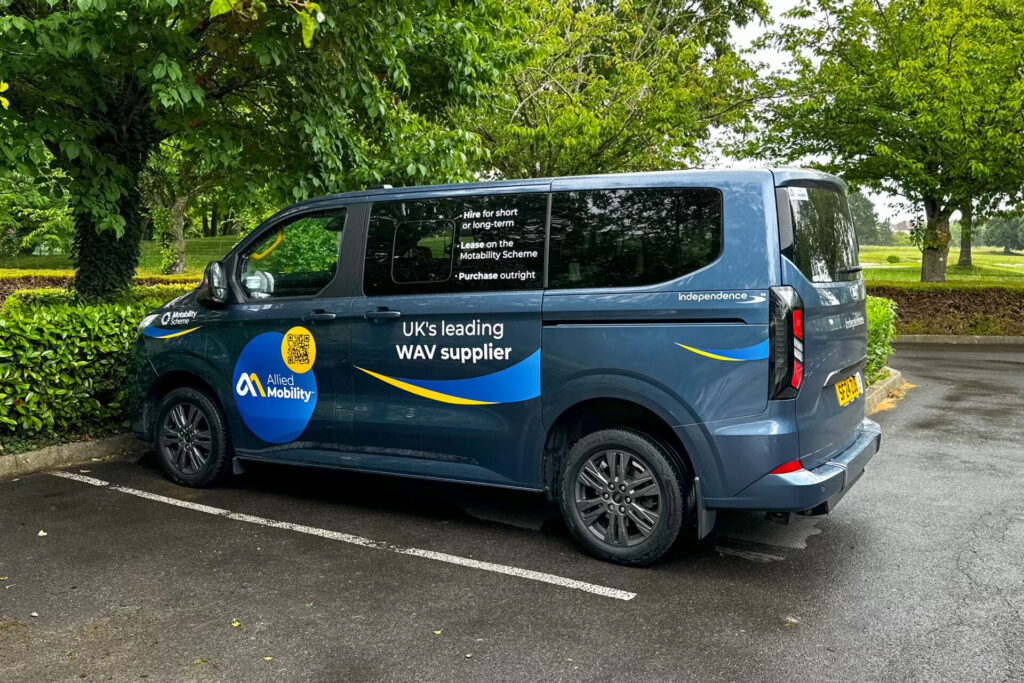 Three quarter back profile photo of a blue-grey 'Allied Mobility' WAV (Wheelchair Accessible Vehicle), parked at a car park surrounded by a nature park filled with trees, plants, & row hedges.