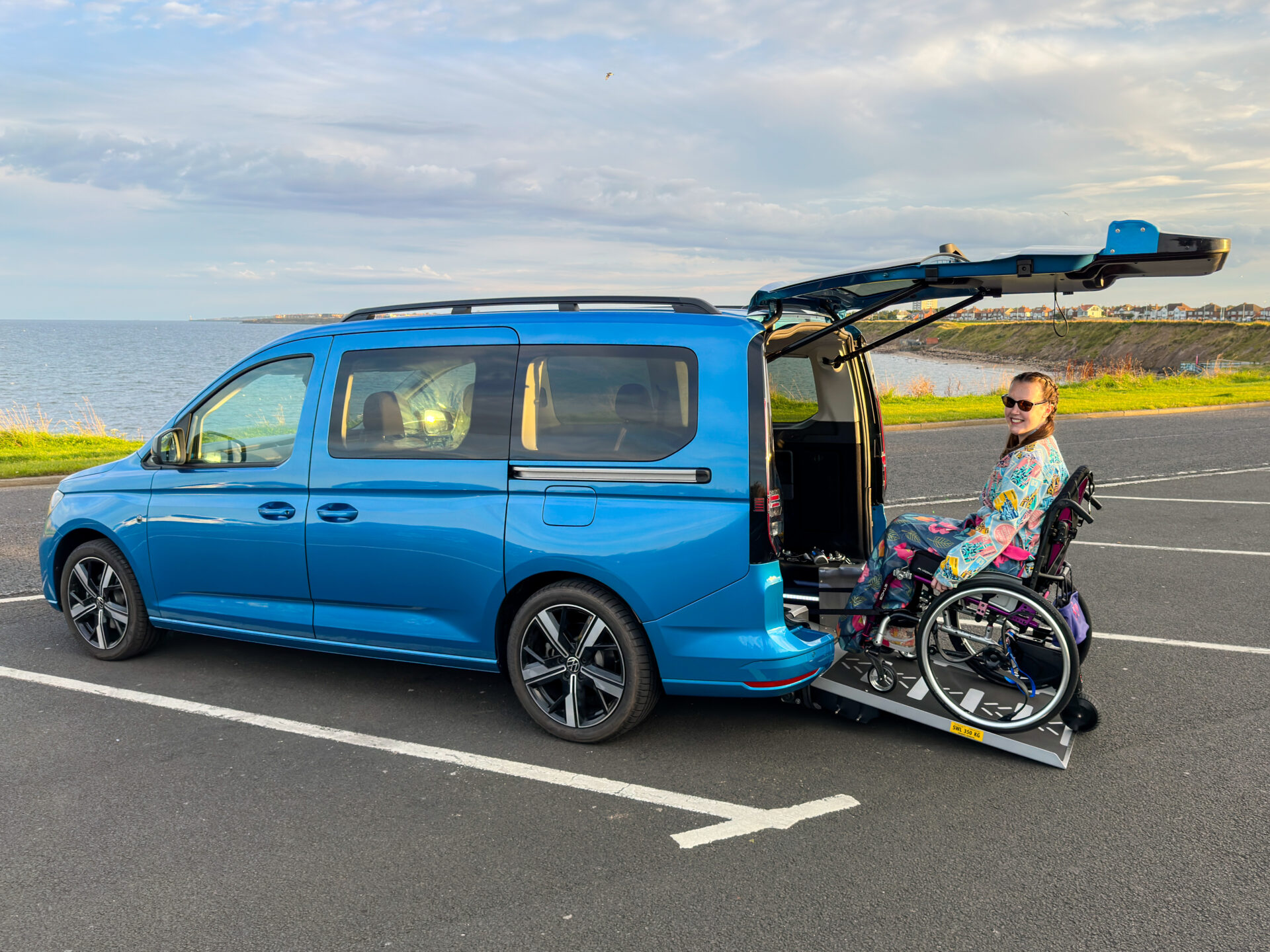 A woman in floral clothing using a rear ramp to access a blue VW Caddy Maxi in a silver manual wheelchair.
