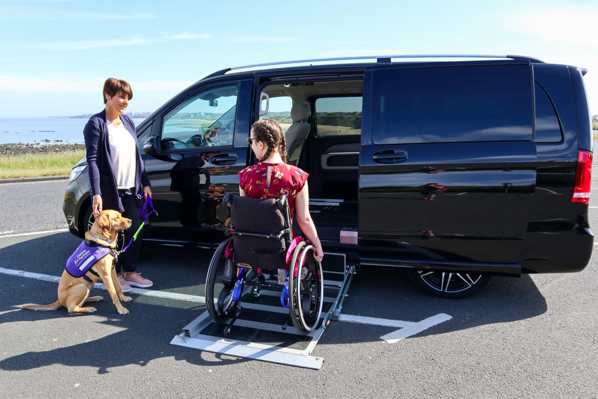 A woman in floral clothing using a side lift to access a black Mercedes V-Class in a pink manual wheelchair.