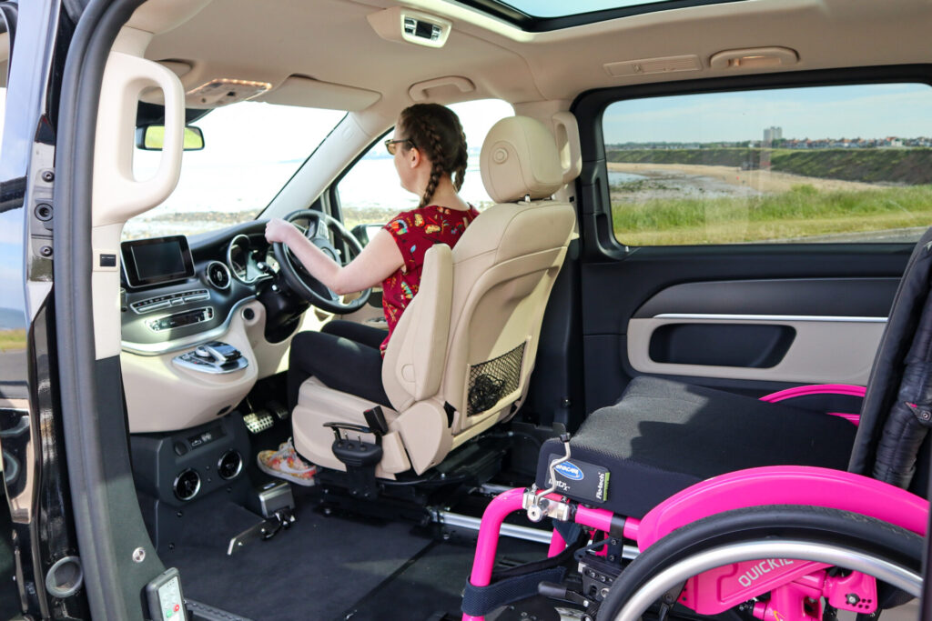 Photo of a woman using a wheelchair accessible vehicle. The woman is sitting in the driver seat, behind which a pink self-propelling wheelchair is stored. The driver's seat is on a set of rails, allowing easy transfer between the seat & the wheelchair.