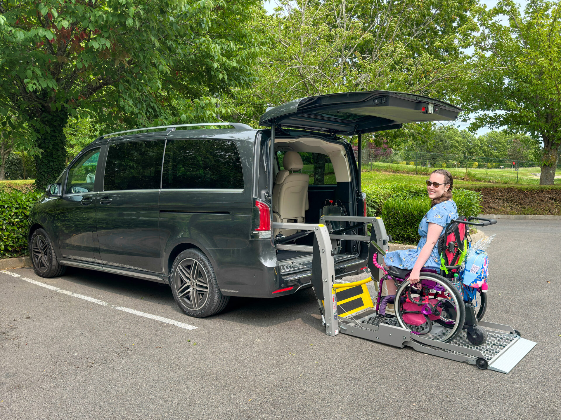 A woman in floral clothing using a rear lift to access a grey Mercedes V-Class in a purple and pink manual wheelchair.