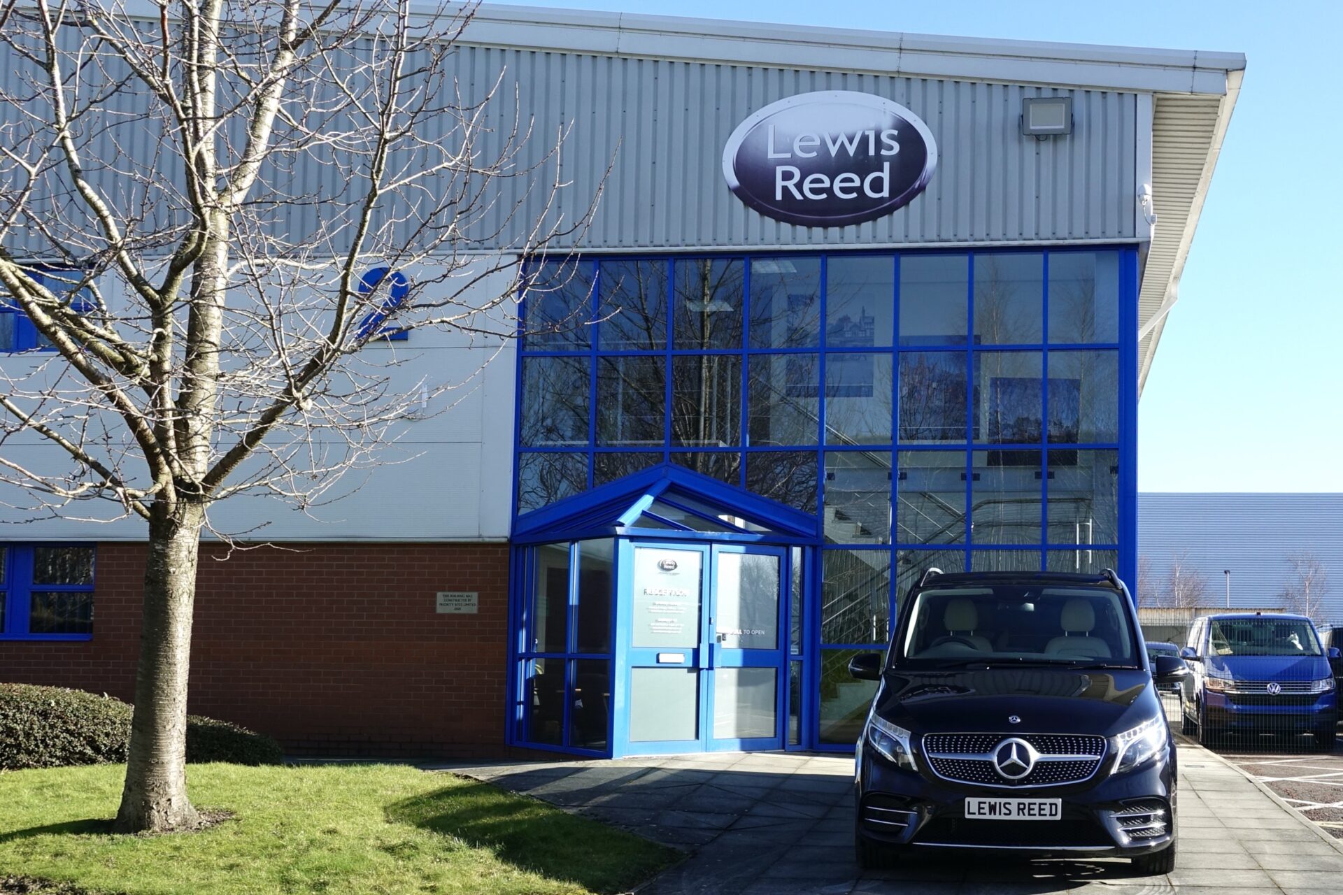 A black Mercedes V-Class parked in front of a building with blue and grey colors, featuring Lewis Reed's logo.