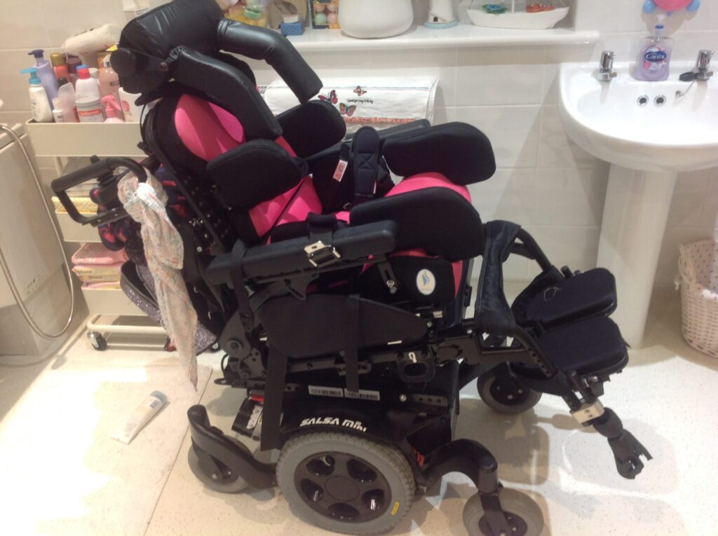 Side profile photo of a 'Quickie Salsa Mini 2' powered wheelchair in a slightly reclined position, in a bathroom, with a black base design, pink coloured seating, arm rests, swing-away foot plates, 4-point harness, lap belt, and a rollercoaster headrest.