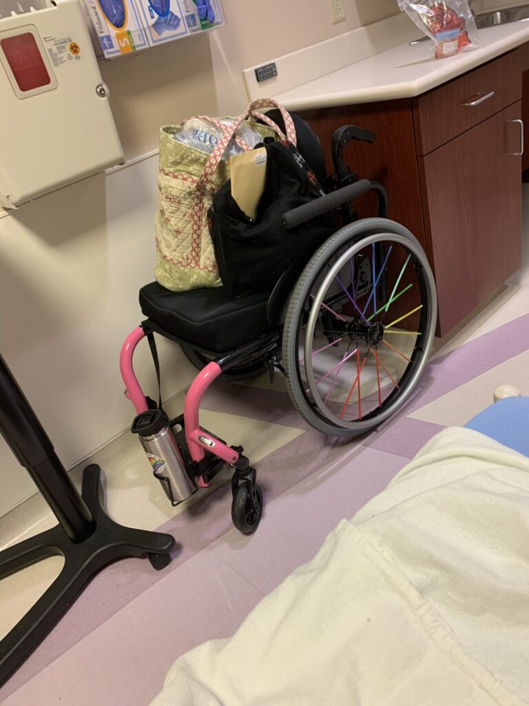 Side profile photo of a 'Ki Mobility Rouge' self-propelling wheelchair, parked next to a desk, with bags stored on the wheelchair seat. The photo is taken from a raised position on a bed (some of which is seen in the foreground of the bottom right corner), which combined with several boxes of disposable gloves and other items fixed to the wall above the wheelchair, indicate this photo was taken in a medical facility, likely as hospital or clinic.