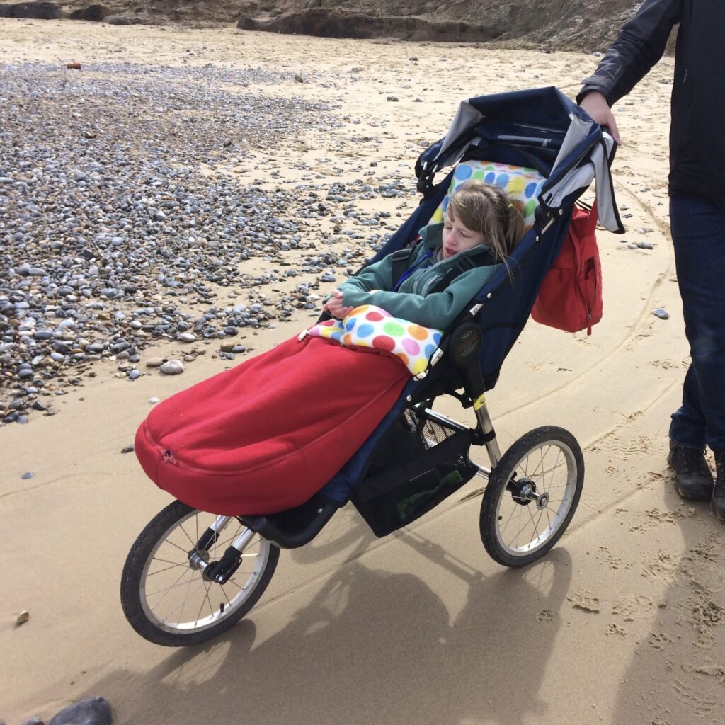 Photo of a child at a pebble beach in an 'Advanced Mobility Independence Buggy', a three wheeled specialist buggy designed to handle rough terrain of places like a rocky, pebbly beach.
