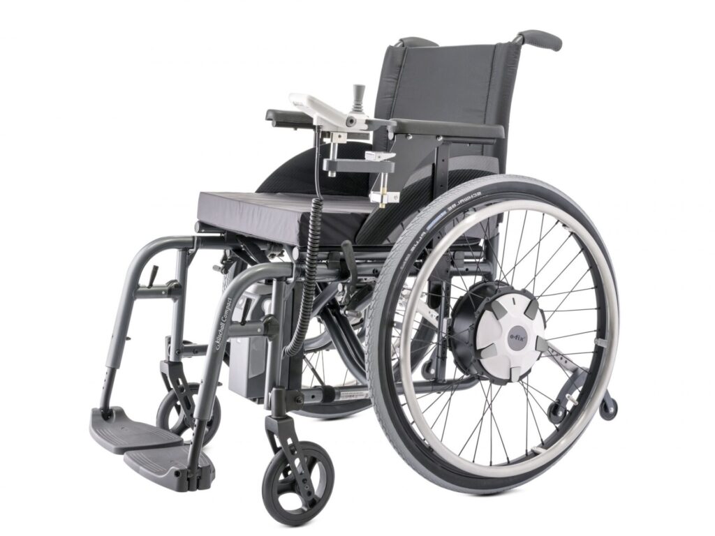 Side profile photo of a wheelchair with 'Alber E-Fix E35' Electric wheelchair attachment wheels & a controller attached. The silver white colour of the attachments identify it as originally separate from the wheelchair, whose frame is a darker, more metallic, grey colour.