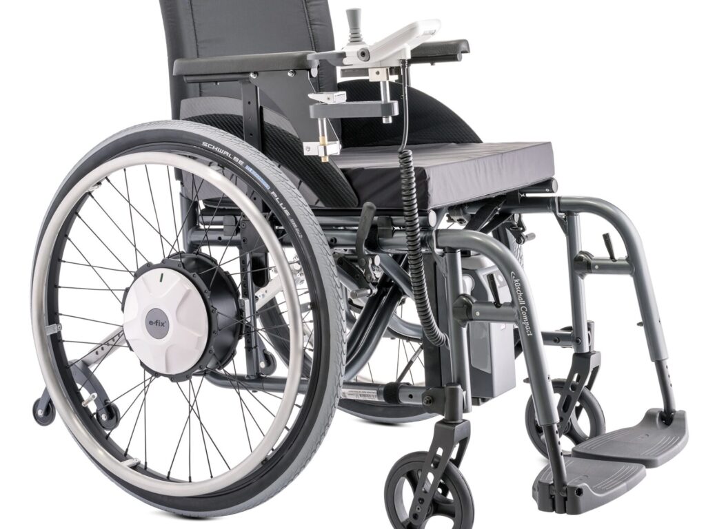 Side profile photo of a wheelchair with a 'Alber E-Fix E35' Electric wheelchair attachment wheels & controller attached. The silver white colour of the attachments identify it as originally separate from the wheelchair, whose frame is a darker, more metallic, grey colour.