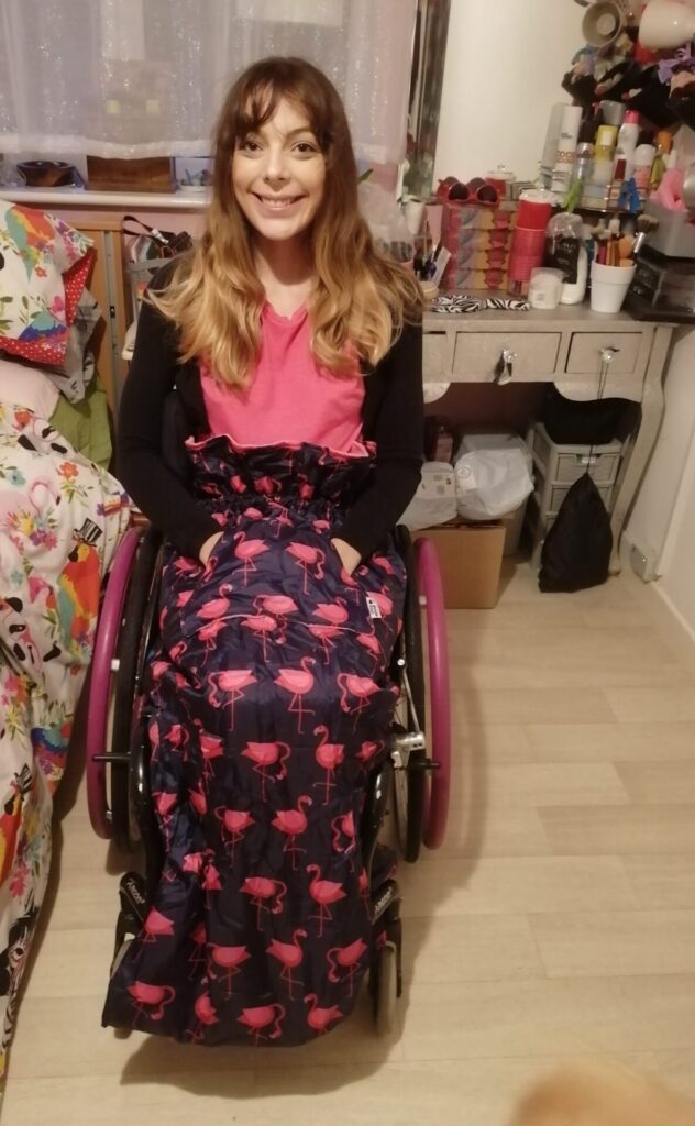 Photo of a smiling woman using a wheelchair drawing attention to the 'Bundle Bean cosy' wheelchair cosy with a repeating flamingo decoration, that she is using.