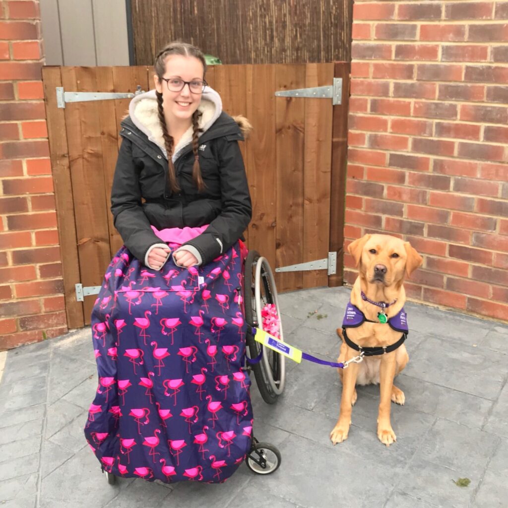 Photo of a female wheelchair user (wrapped up warm in a black winter coat, fleeced hoodie, & a purple BundleBean cosy, decorated with flamingos) with her 'Canine partner' dog (identified by their purple coat), posing for a photo.