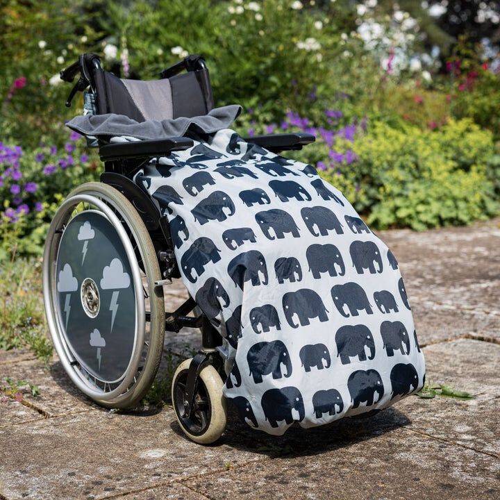 Photo of a self-propelling wheelchair with a white 'BundleBean' wheelchair cosy, decorated with a repeating rows of black stencil elephants, attached to it. Four 'white cloud with lightning' patterns on a black background decorate the wheelchair wheel's spoke guards.