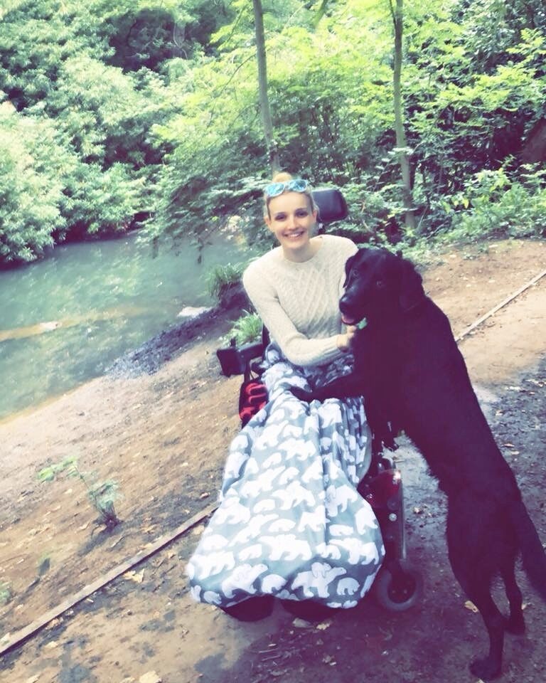 Photo of a woman using an electric wheelchair, wearing a black & white 'BundleBean' wheelchair cosy with a stencil elephant pattern, and her large black dog, which has its front paws on the woman's lap, as turns around to look at the camera, posing for a photo at the top of steep bank over looking a shallow stream.