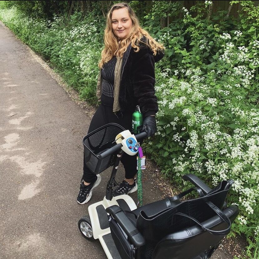 Photo of a blonde haired woman dressed all in black, standing with a 'Clearwell Aguna' mobility scooter in front of her, holding on to its right steering handle on a gravely path. In the background a large number of white flowers, and small woody plants can be seen.