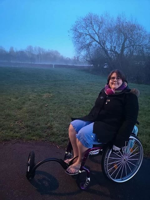 Photo of a woman using a self-propelling wheelchair, with a 'FreeWheel' attachment connected to the footplate, on a footpath in a park. The woman's scarf and thick black coat, contrast with he blue shorts & sandals, which in conjunction with the the low light, and mist in the far background, suggest this photo was taken very early in the morning on a summer day.