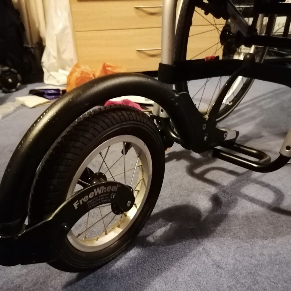 Photo of a 'FreeWheel' front wheel attachment, connected to the front of a wheelchair by a clamp that clips onto the wheelchair's footplate.