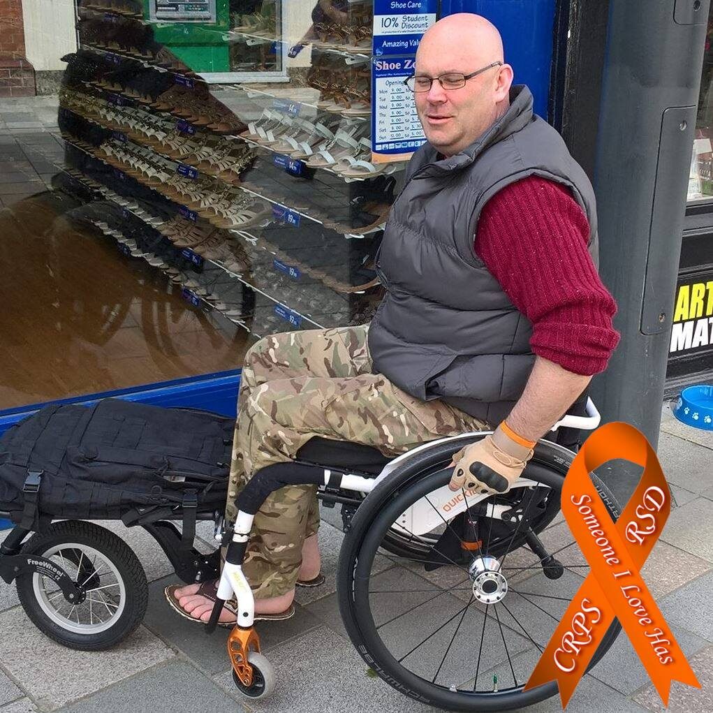 Photo of a man using a self-propelling wheelchair, with a 'FreeWheel & Rack' attached to the front of the wheelchair, allowing the man's bag to be easily transported.