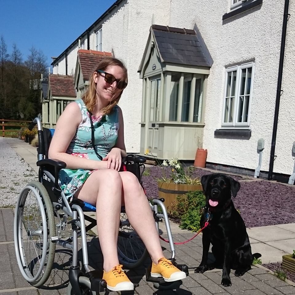 Photo of a smiling woman using a 'Excel G-Lite Pro' folding frame, self-propelling wheelchair, posing for a photo in front of a row of house with her small black dog wearing a red leash.