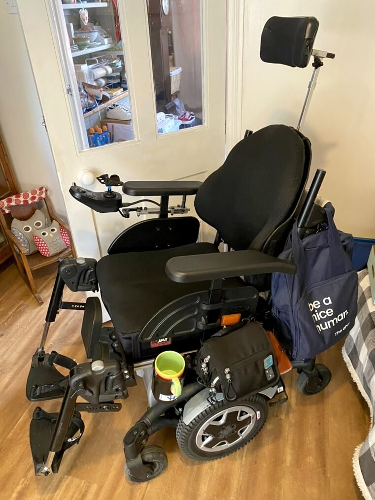 Photo of a an electric wheelchair with an attached 'Handicup' cupholder just visible to the side & below the left hand wheelchair arm. Several bags hanging on parts of the wheelchair, demonstrate the the large number of things the user needs on a daily basis, which highlights the immense value of a dedicated space for beverages that the cup holder provides.