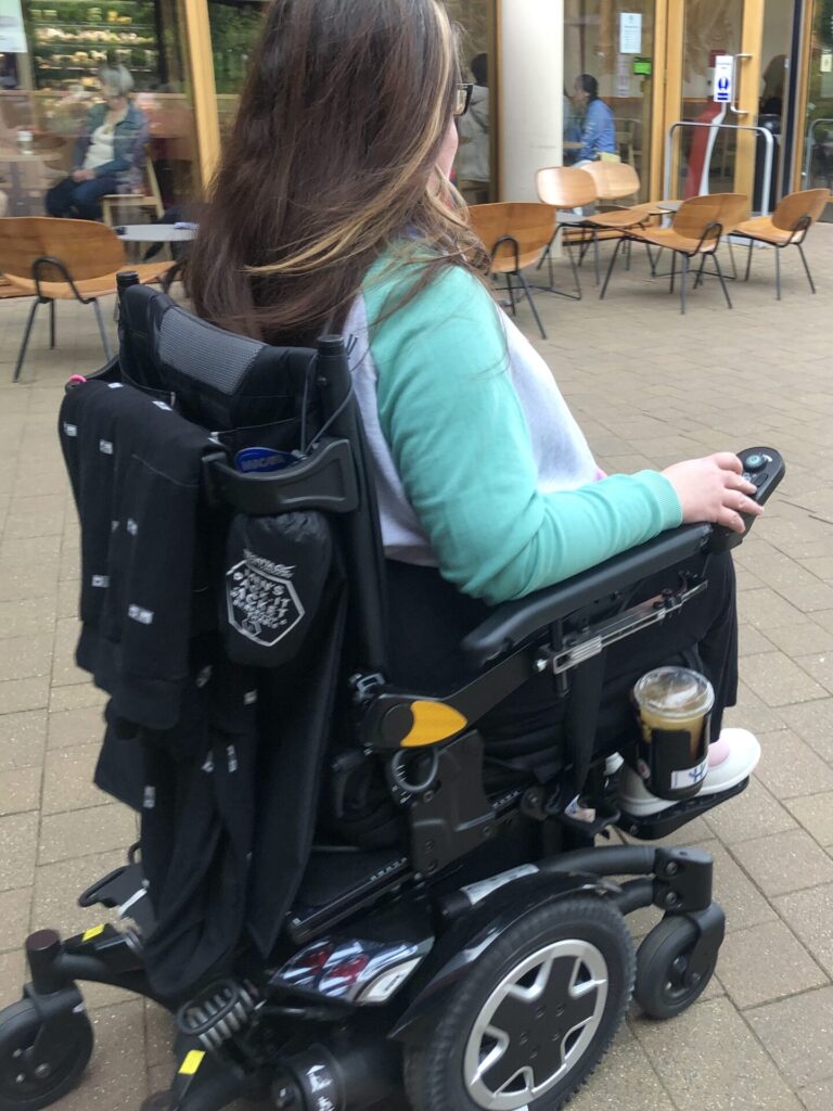 Side profile photo of a woman using a wheelchair with a cup holder attachment connected between the wheelchair cushion and the seat base.