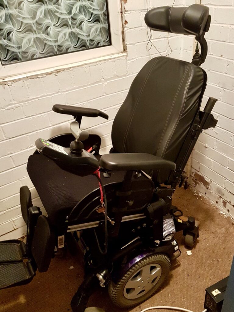Photo of an 'Invacare TDX SP2 NB' electric wheelchair. The backrest has been put into a slightly reclined position, while the wheelchair is parked in the corner of a room.