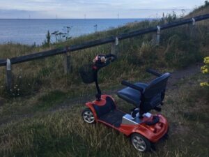 Photo of a 'Kymco Mini LS' Mobility Scooter, with its distinct orange metal frame work, parked on a overgrown cliffside path, a wooden guardrail bordering the path, separating it from the ocean view in the background, where a line of wind turbines are visible.