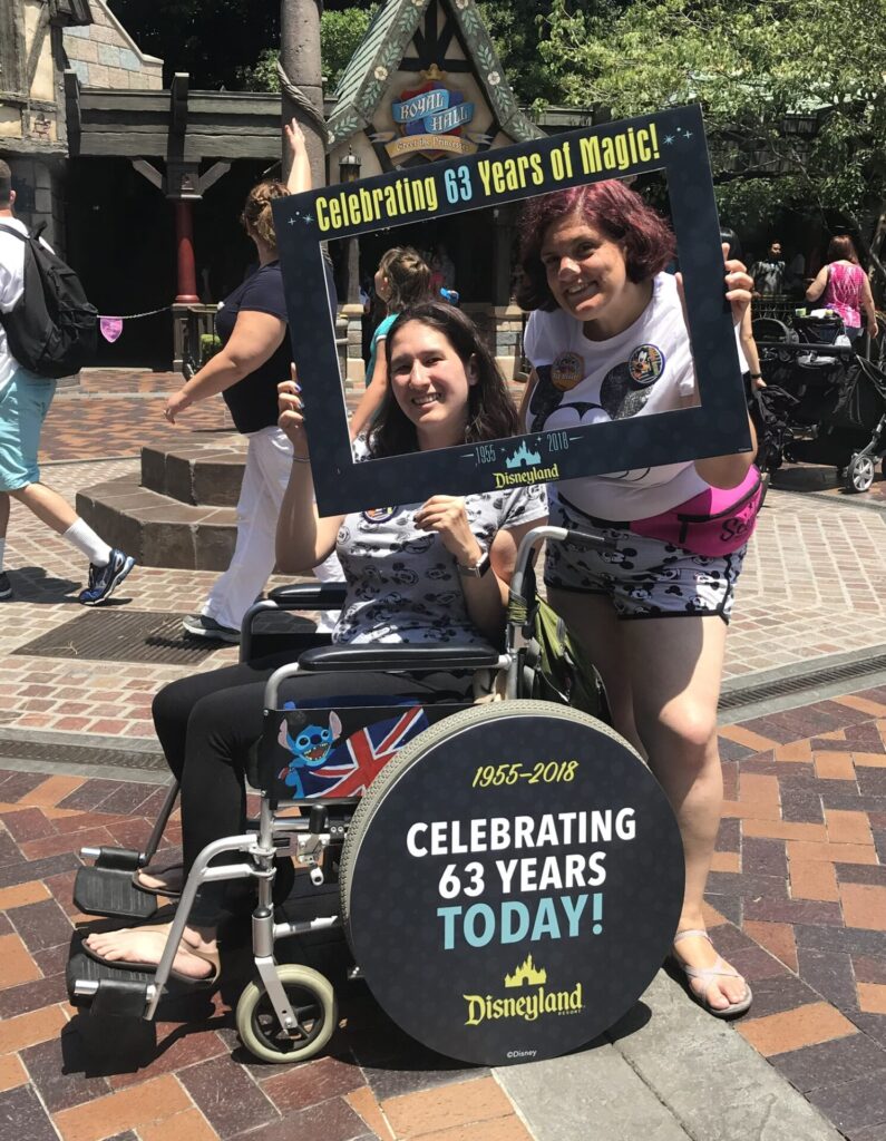 Photo of two women, one of whom is using a wheelchair, posing while using a frame to highlight their smiling faces, that reads 'Celebrating 63 Years of Magic' at the top & 'Disneyland' at the bottom. A cover on the cap of a wheel of the woman's wheelchair says the same information, as well as '1955-2018' & 'Today!'