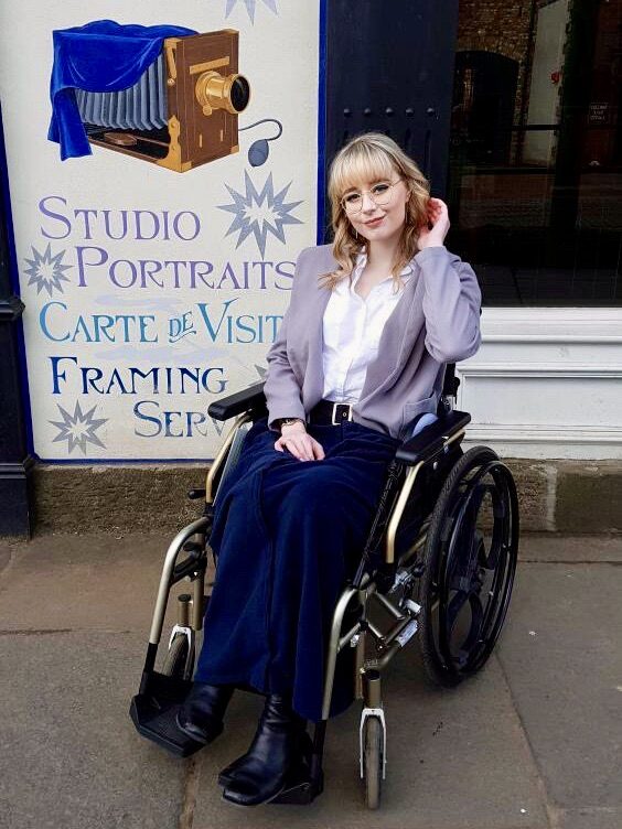 A bespectacled female wheelchair user, with 'Loopwheels Classic' installed on their wheelchair, in smart casual business attire, wearing a navy blue blanket, posing for a photo in front of a large sign advertising 'Studio Portraits' with an stylized drawing of an 19th century studio camera.