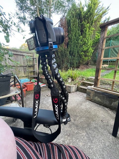 Photo of a 'Meru Flexzi 3' three-stranded adjustable device holder, holding a camera in place on top of the lefthand arm of a wheelchair.