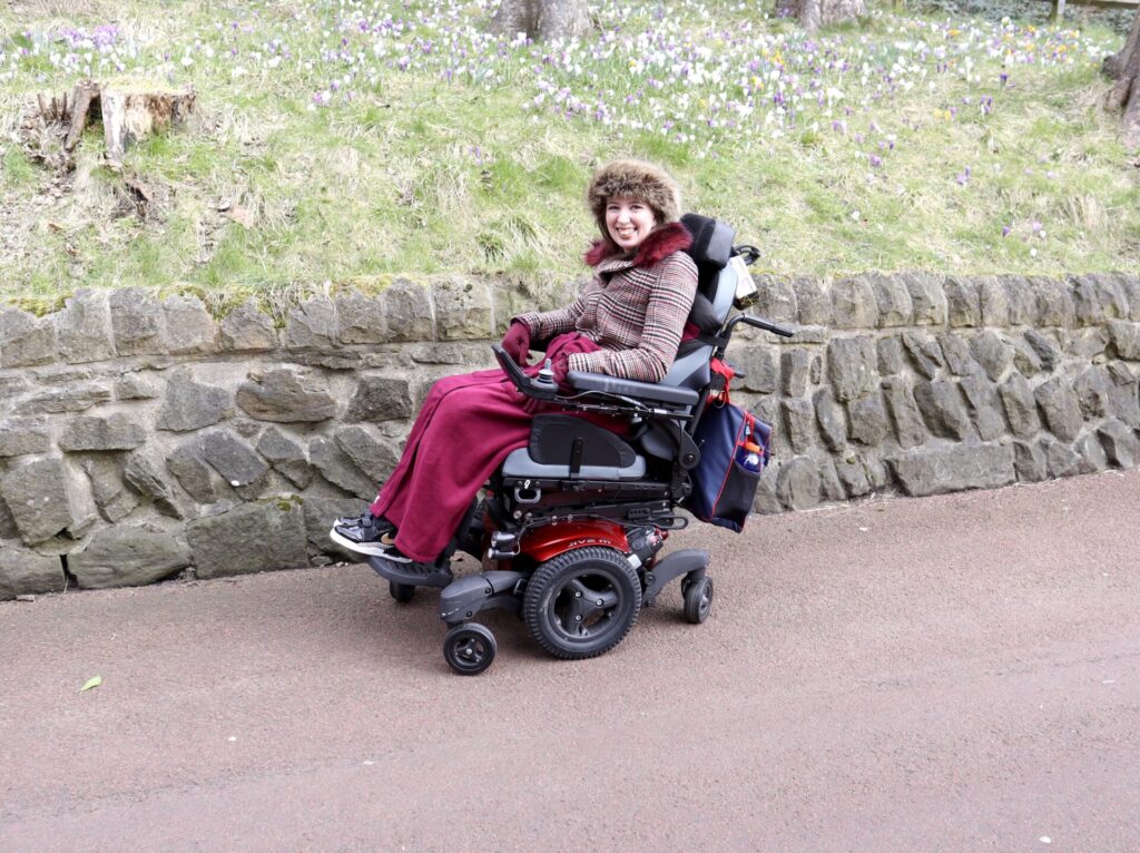 Photo of a smiling woman in a electric wheelchair, on a paved path in a garden park. The fur-trimmed hat, large striped red coat, & pink 'NiCosy Blanket' leg covering she is wearing, indicates it is a cold day. In the background a carved stone wall, & a hill covered in white & purple flowers can be seen.