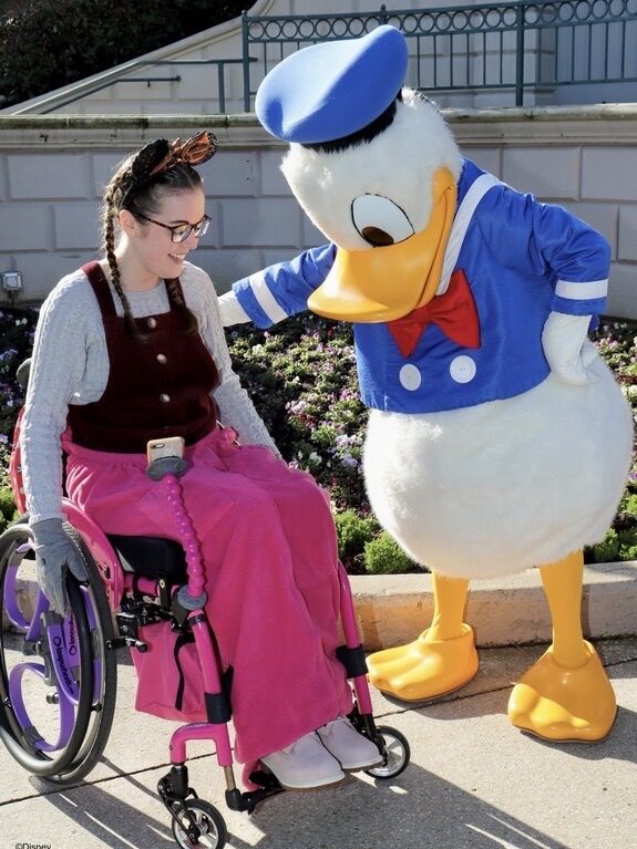 Photo of a female wheelchair user, wearing a pink NiCosy wheelchair blanket. The "Donald Duck" mascot standing next to her, indicates that this photo was taken at one of the "Disneyland" theme parks.