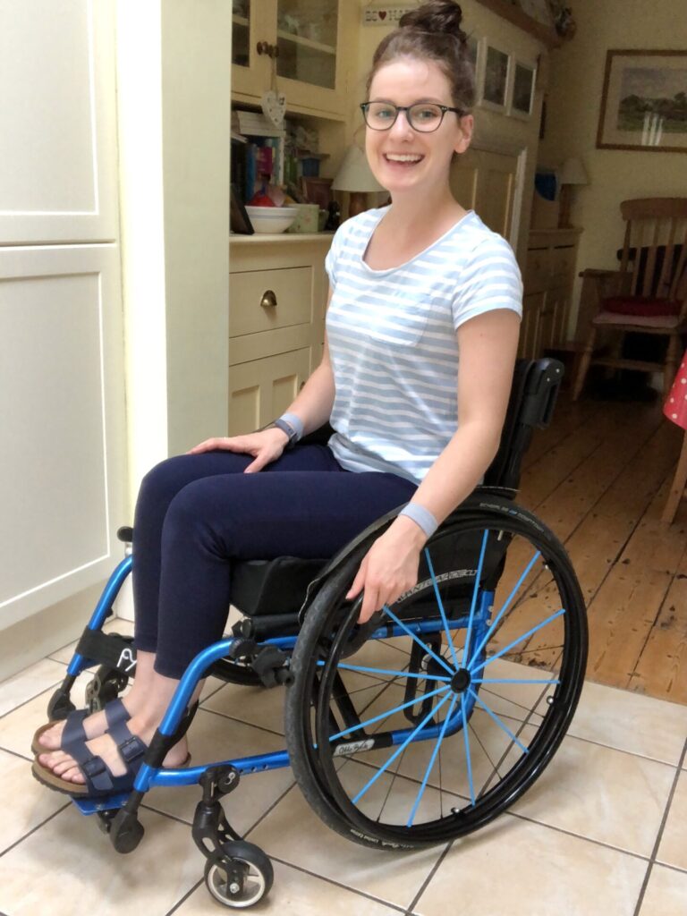 Three quarter profile photo of a smiling bespectacled woman using an 'Ottobock Avantgarde CLT' foldable self-propelling wheelchair on a floor of large panels.