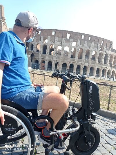 Photo of a man using a 'Pandhora EVO' wheelchair with a 'Pandhora P3' bike style electric attachment connected to the front of the wheelchair, posing in front of a colosseum.